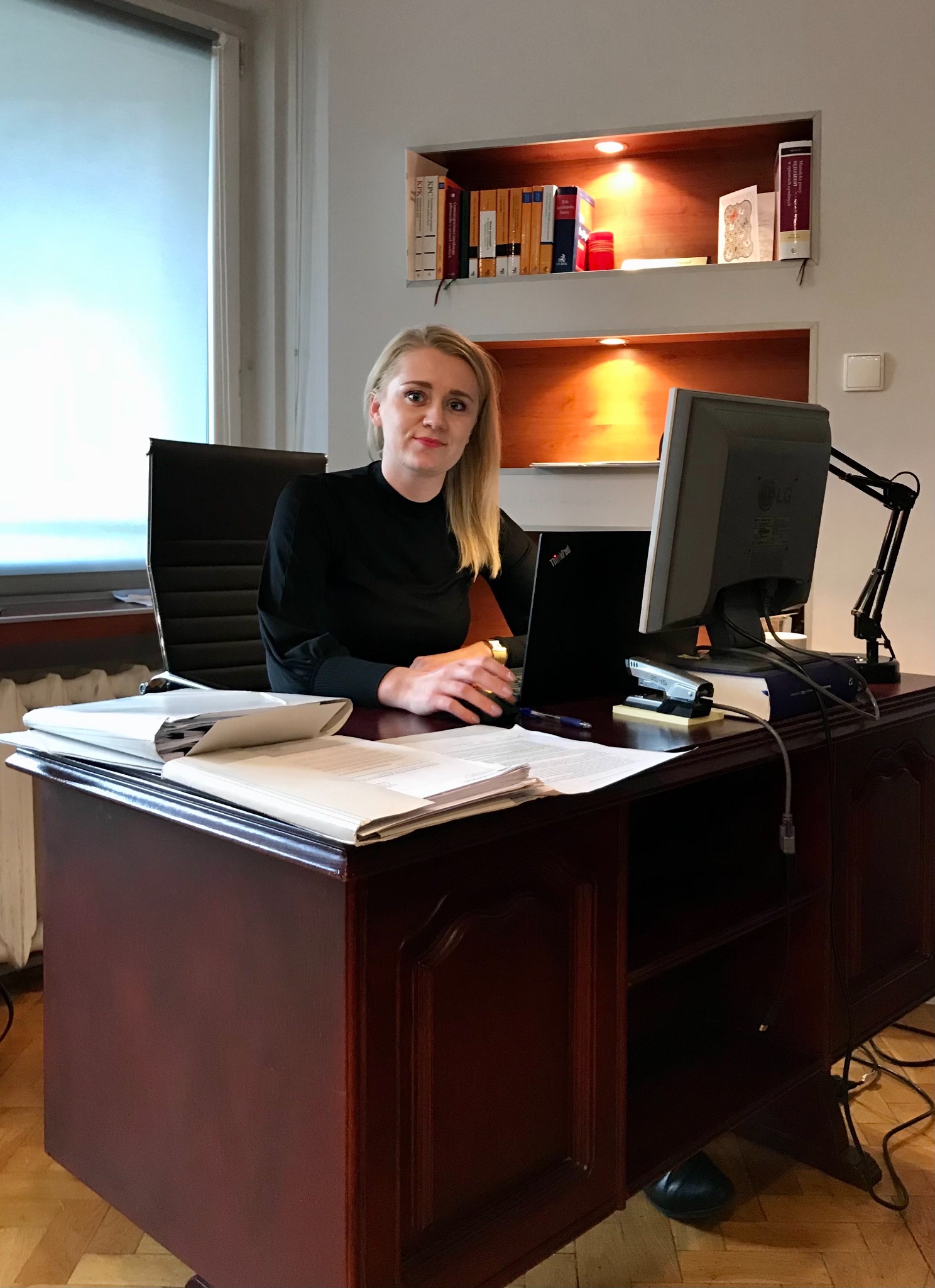 Kobieta w czarnej bluzce siedzi za biurkiem z laptopem i stosem dokumentów, w tle regał z książkami i lampka biurkowa.