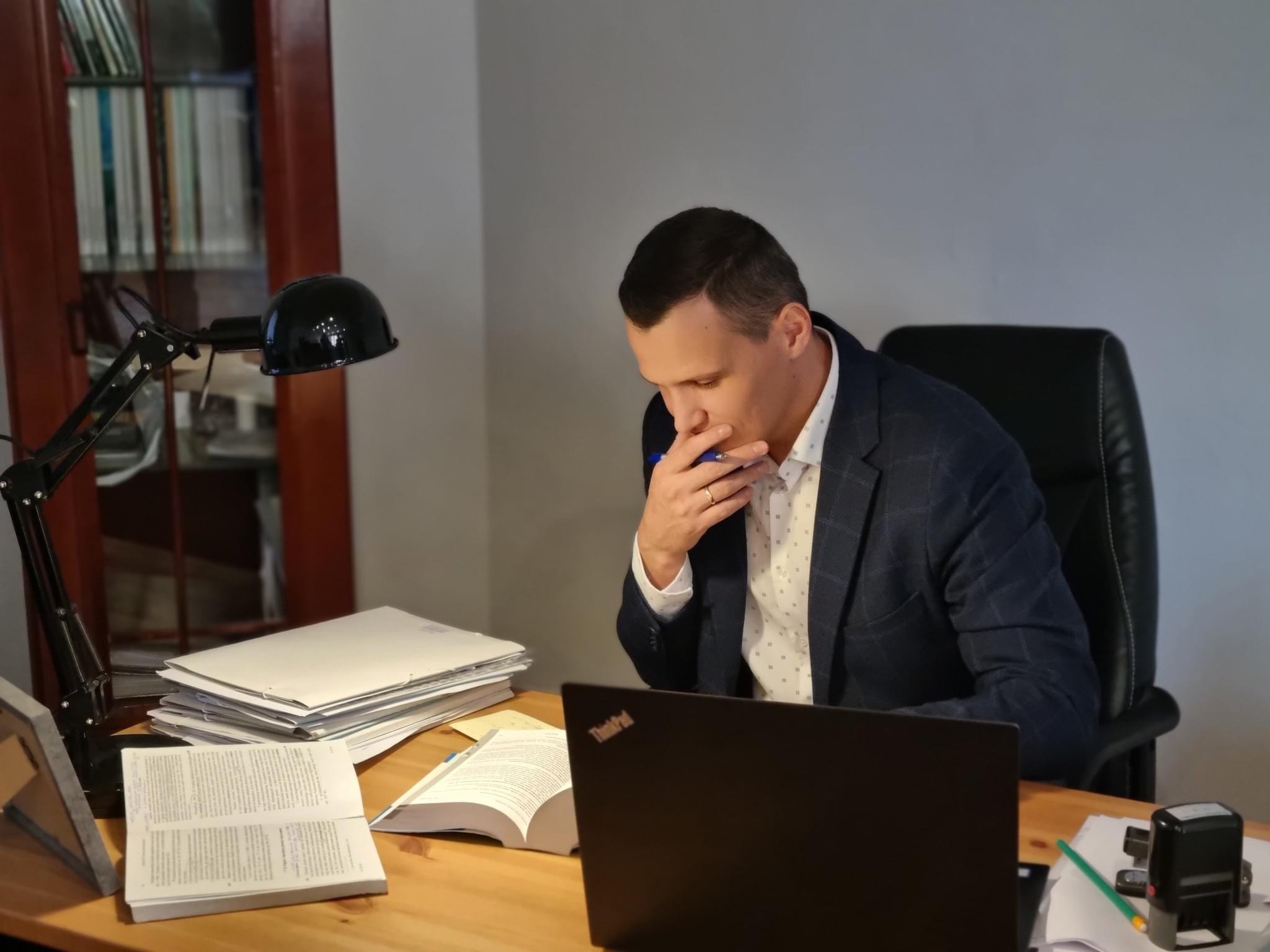 Mężczyzna w garniturze siedzi przy biurku z laptopem i otwartą książką, zamyślony trzyma długopis przy ustach. Na biurku sterta dokumentów i lampka.