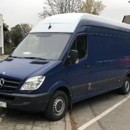Niebieski bus Mercedes Sprinter z białym dachem na tle budynków i drzew, zaparkowany na kostce brukowej. Widoczna tablica rejestracyjna.
