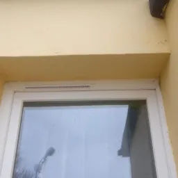 Fragment okna z białymi ramami i widoczną kratką wentylacyjną, osadzonego w żółtej ścianie budynku z widocznym fragmentem elewacji i narożnikiem.