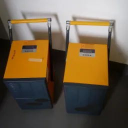 Dwa identyczne, przenośne rekuperatory w obudowach żółto-szarych, z żółtymi uchwytami, stojące na szarej podłodze przy białej ścianie.