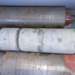 Rdzenie z betonu wycięte wiertnicą diamentową, ułożone w szarej kuwecie z parą roboczych rękawic i metalową złączką.