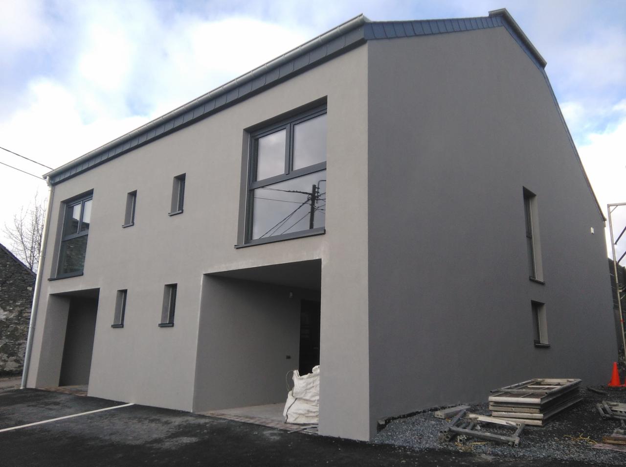 Nowoczesny, dwukondygnacyjny dom z szarą elewacją i ciemnymi oknami, widok z zewnątrz, z workiem materiałów budowlanych pod wjazdem do garażu i stertą ram okiennych obok.