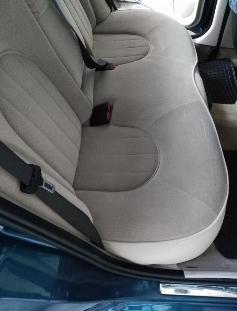 Jasna, welurowa tapicerka tylnej kanapy w samochodzie, widoczne pasy bezpieczeństwa i fragment progu w kolorze ciemnego turkusu.