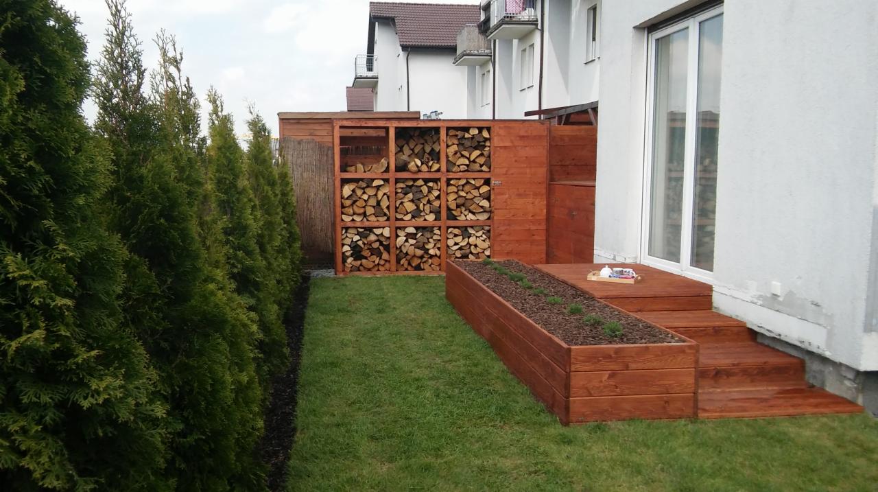 Nowoczesny ogród w Gnieźnie z drewnianym podestem, rabatą i regałem na drewno, oddzielony od sąsiada szpalerem tuj.