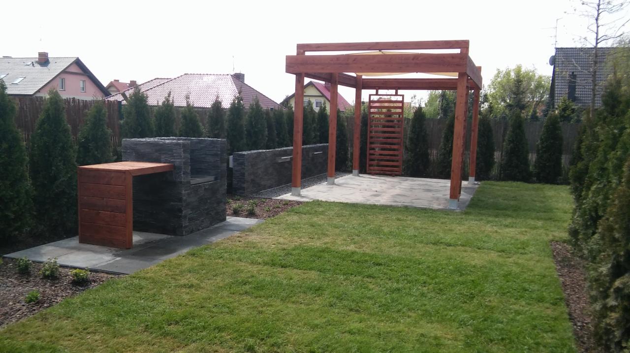 Nowoczesny ogród z murowanym grillem z kamienia łupanego, drewnianą pergolą z zacienieniem i zielonym trawnikiem, całość otoczona rzędem wysokich, gęstych tuj.