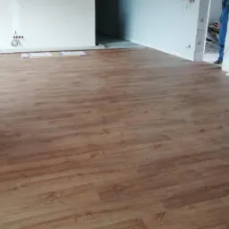 Ułożona podłoga z paneli imitujących drewno w jasnym, prawie pustym pokoju w trakcie remontu, widoczne puszki elektryczne na ścianie i fragment osoby w jeansach.