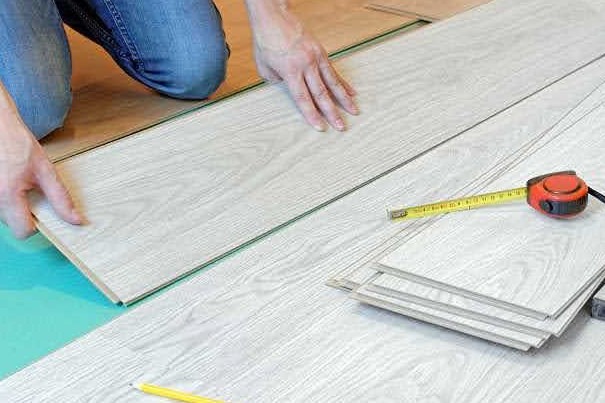 Układanie paneli podłogowych w jasnym odcieniu drewna, widoczna miarka i ołówek, osoba dociska panel do podłoża.