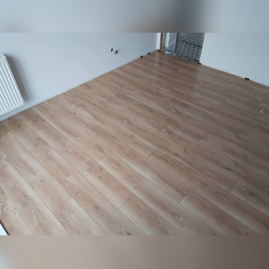 Ułożone panele podłogowe w odcieniach jasnego drewna, widoczny fragment grzejnika i biała ściana z gniazdkiem elektrycznym, w tle wejście do innego pomieszczenia z szarą podłogą w romby.