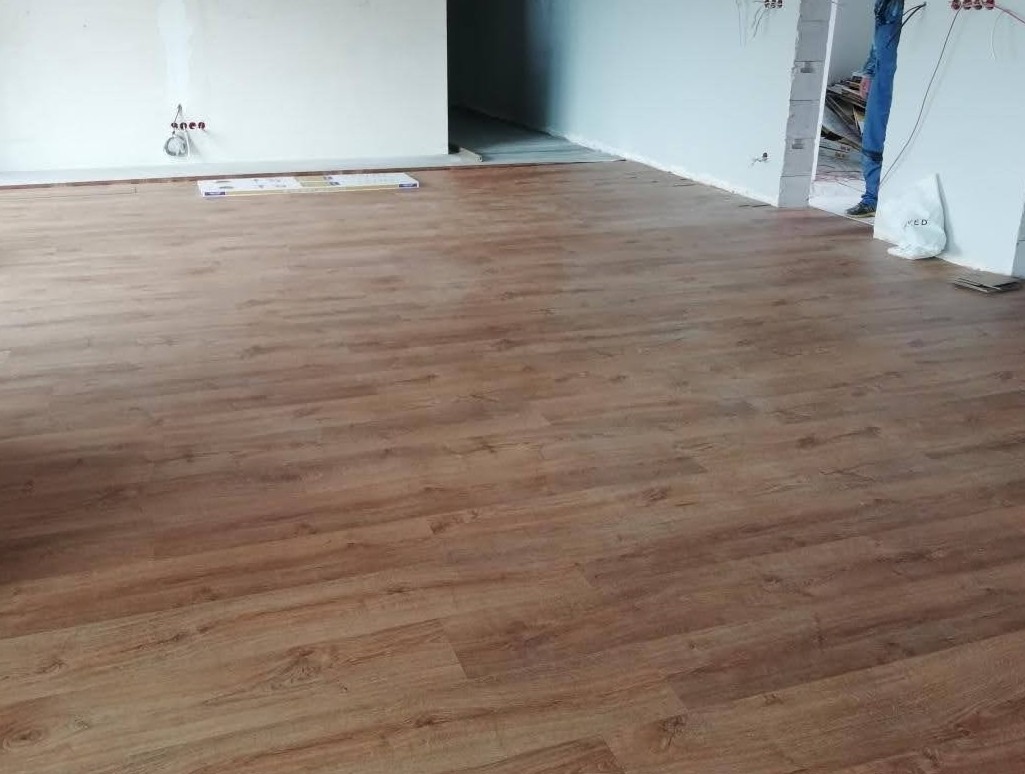 Ułożona podłoga z paneli imitujących drewno w jasnym, prawie pustym pokoju w trakcie remontu, widoczne puszki elektryczne na ścianie i fragment osoby w jeansach.