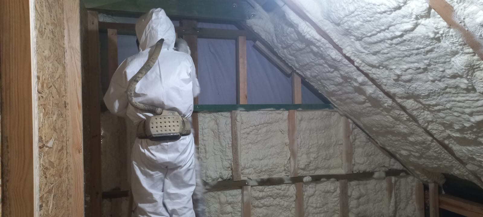 Pracownik w kombinezonie ochronnym aplikuje izolację piankową na poddaszu, widoczne drewniane elementy konstrukcyjne i świeżo nałożona pianka o nieregularnej fakturze.