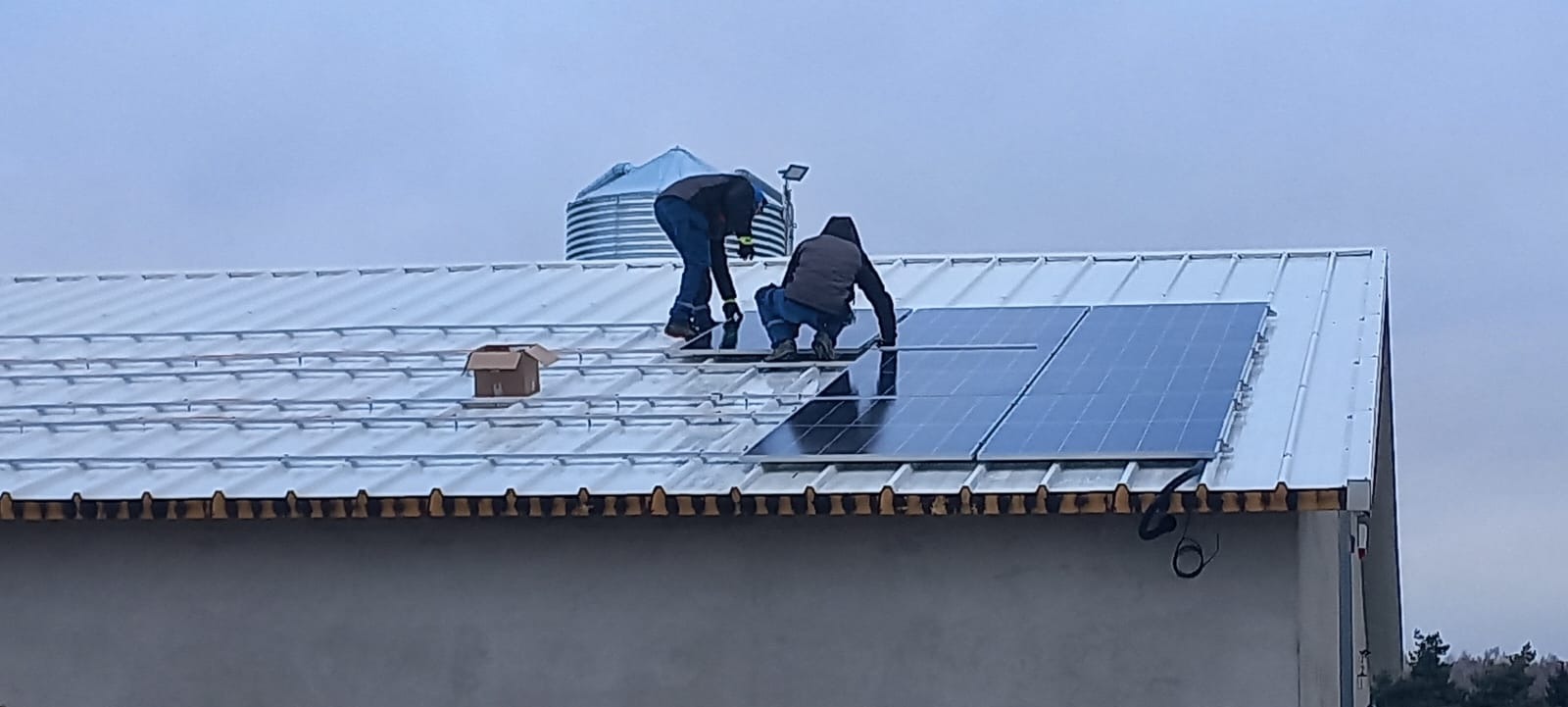 Montaż paneli słonecznych na metalowym dachu budynku gospodarczego; widoczni dwaj monterzy w roboczych ubraniach, kartonowe pudło i silos w tle.