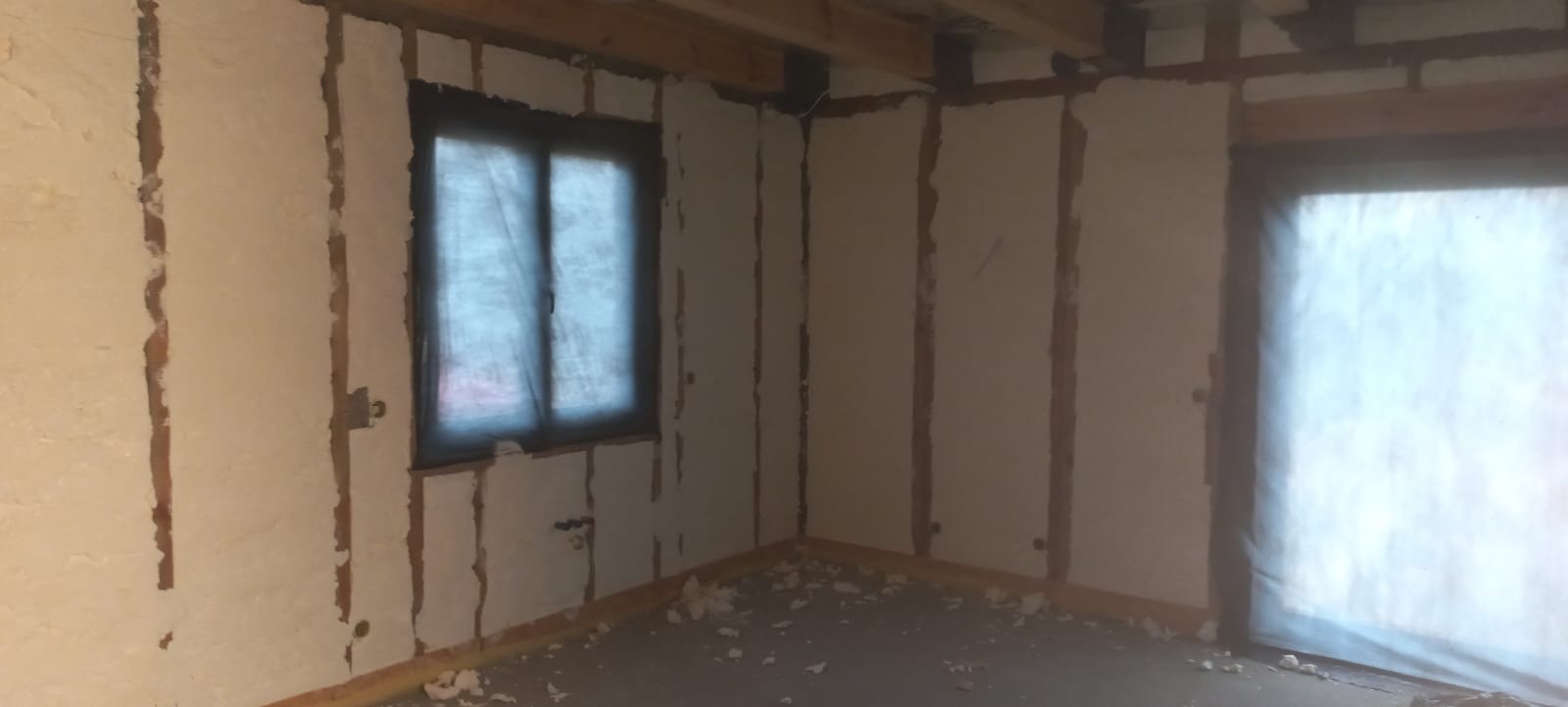 Wnętrze domu w trakcie prac ociepleniowych: ściany pokryte pianką, widoczne drewniane elementy konstrukcyjne, okno zabezpieczone folią, fragment podłogi z resztkami pianki.