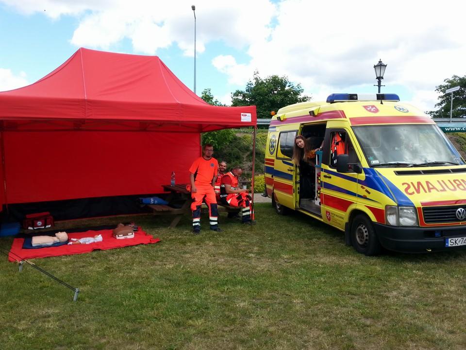Prezentacja z zakresu pierwszej pomocy: ambulans, namiot, manekiny do ćwiczeń resuscytacji oraz personel w pomarańczowych uniformach.