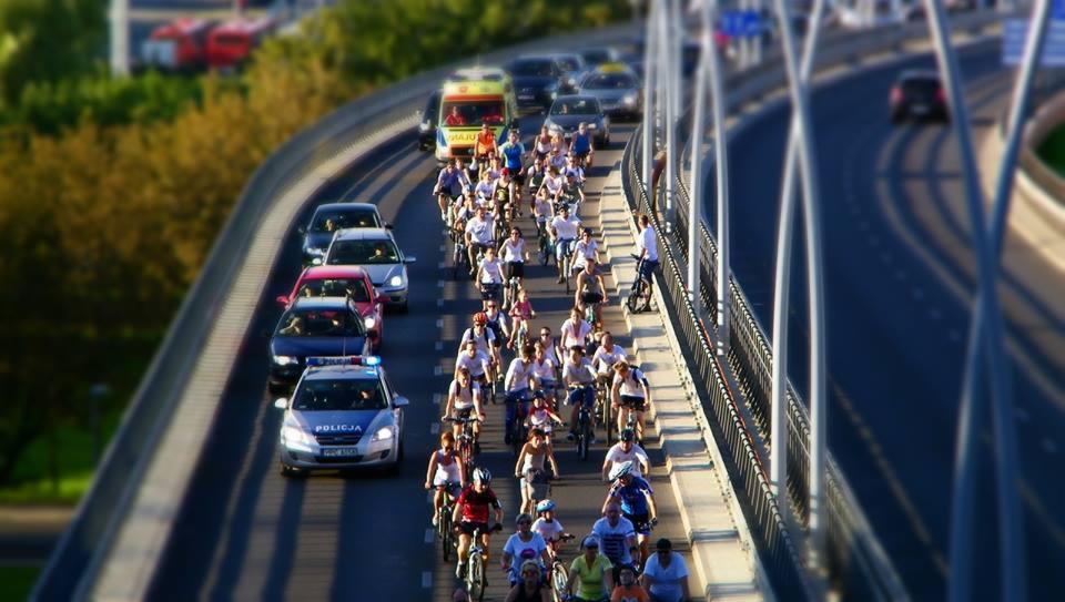 Kolumna rowerzystów eskortowana przez policję i ambulans przejeżdża przez most w słoneczny dzień.