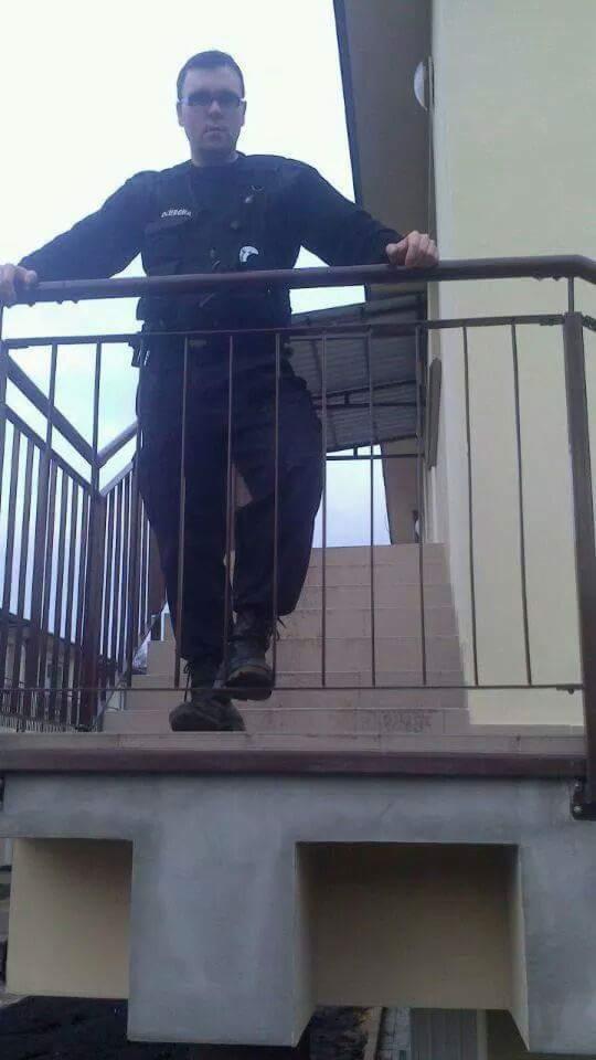 Mężczyzna w czarnym uniformie i okularach, stojący na schodach z metalową barierką, widok z dołu.