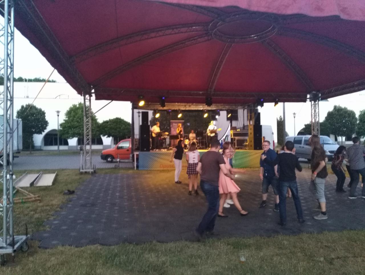 Zespół muzyczny grający na scenie pod zadaszeniem, przed sceną tańczą ludzie, w tle budynek i zaparkowane samochody.