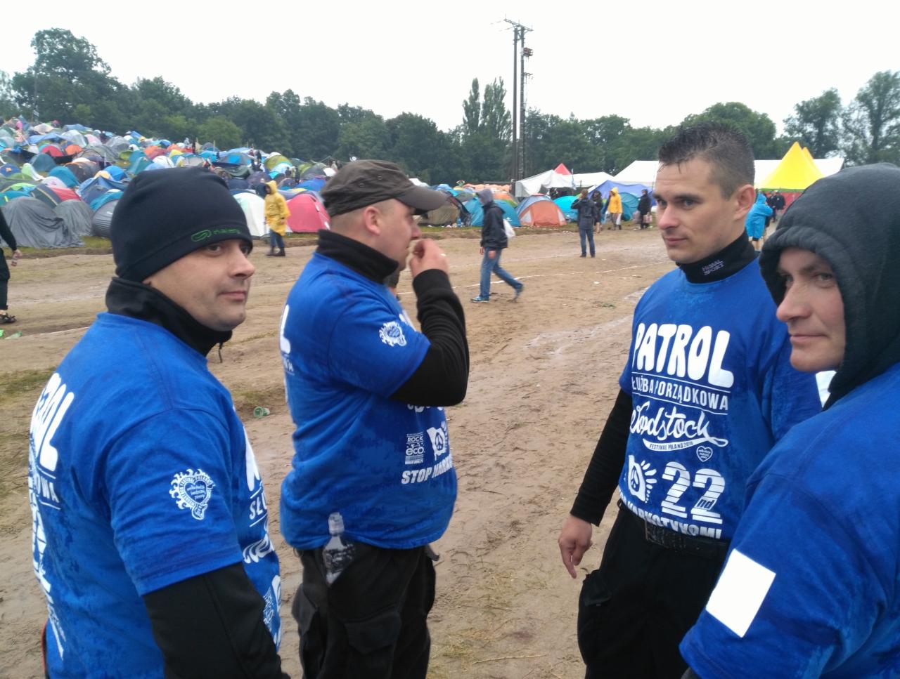 Czterech mężczyzn w niebieskich koszulkach z napisem 'Patrol' na tle pola namiotowego, prawdopodobnie podczas zabezpieczania imprezy masowej. W tle widoczne liczne namioty i osoby. Zdjęcie wykonane...