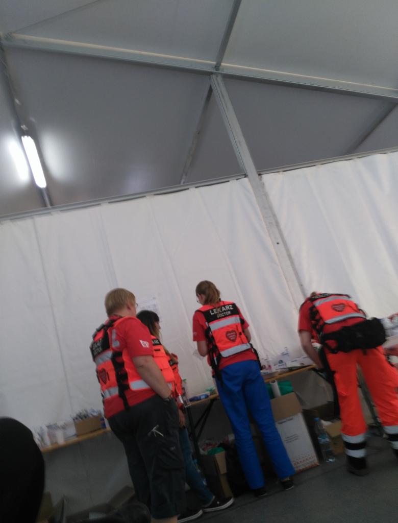 Grupa ratowników medycznych w czerwonych koszulkach i kamizelkach odblaskowych z napisem 'Lekarz' wewnątrz namiotu z białymi ścianami, podczas przygotowań lub odpoczynku.