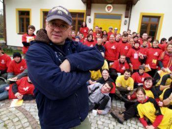 Mężczyzna w okularach i czapce na pierwszym planie, za nim grupa osób w czerwonych i żółtych koszulkach przed budynkiem z białymi ścianami i brązowym dachem. Osoby siedzą na ziemi. W tle drzewa.