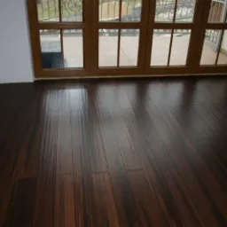 Ciemna, lakierowana podłoga z desek ułożona wzdłuż dużych, brązowych okien z widokiem na balkon z metalową balustradą.