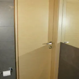 Jasne, drewnopodobne drzwi wewnętrzne z chromowaną klamką, zamontowane w łazience z szarą płytką ceramiczną i odbiciem w lustrze.