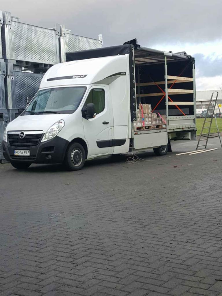 Biały bus z otwartą plandeką, załadowany paletami z kartonami, na parkingu z szarej kostki brukowej, drabina oparta o tył pojazdu.