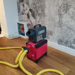 Profesjonalny osuszacz powietrza marki KoronaOzon.pl, z podłączonymi żółtymi wężami, ustawiony na parkiecie w pomieszczeniu z ozdobną tapetą i widocznymi śladami wilgoci przy listwie przypodłogowej.