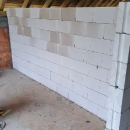 Wznoszenie ściany działowej z białych bloczków na poddaszu, widoczne drewniane krokwie, częściowo otynkowana ściana z cegły, narzędzia i akcesoria budowlane na podłodze.