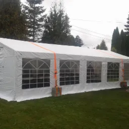 Biały namiot imprezowy z oknami, zabezpieczony pomarańczowymi pasami transportowymi, ustawiony na trawie w ogrodzie.