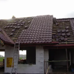 Budowa domu jednorodzinnego w trakcie, widoczny częściowo pokryty dach z brązowej dachówki, rusztowanie i drabina oparta o ścianę.