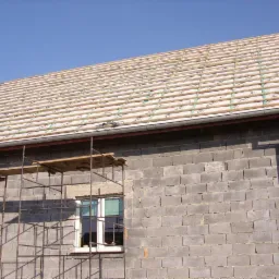 Budynek w trakcie prac dekarskich: widok na szkielet dachu z łatami i membraną dachową, rusztowanie przy ścianie z szarego betonu komórkowego i okno z białą ramą.