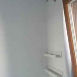 Białe, świeżo pomalowane wnętrze pokoju z dwoma białymi półkami na ścianie, karniszem i fragmentem okna z drewnianą ramą.