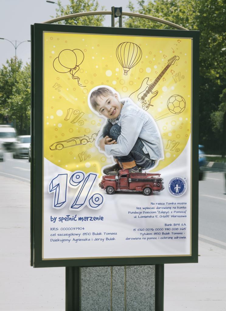 Billboard reklamowy na ulicy z ilustracją chłopca, czerwonej zabawki-ciężarówki i napisem '1% by spełnić marzenie' oraz danymi fundacji i banku, zachęcający do przekazywania 1% podatku.