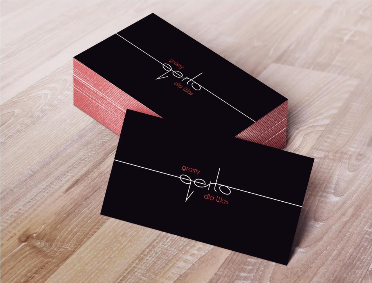 Stos czarnych wizytówek z różowymi brzegami i białym logo 'gramy perto dla Was' leżący na drewnianym stole, jedna wizytówka leży płasko obok.