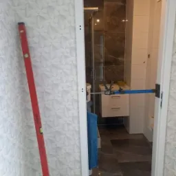 Montaż ościeżnicy w łazience z widocznym poziomem, narzędziami i fragmentem wykończonej łazienki z kabiną prysznicową w tle.