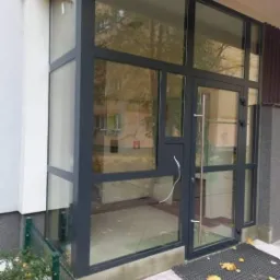 Szklana zabudowa wejścia do budynku z szarymi ramami, widoczny fragment chodnika z opadłymi liśćmi i wycieraczka.