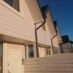 Szereg identycznych, pomalowanych na biało domów z drewnianą elewacją, ciemnymi dachami i rynnami, widzianych z perspektywy żabiej zza niskiego, białego płotku.