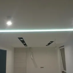Nowoczesne oświetlenie sufitowe z listwą LED i reflektorami punktowymi w nowo wykończonym, pustym pomieszczeniu z białymi ścianami i widocznymi niedoskonałościami w postaci przewodów i otworów.
