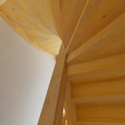 Wewnętrzny widok spiralnych schodów z jasnego drewna, z widoczną strukturą słojów, wspartych na solidnym drewnianym słupie, na tle białej ściany.