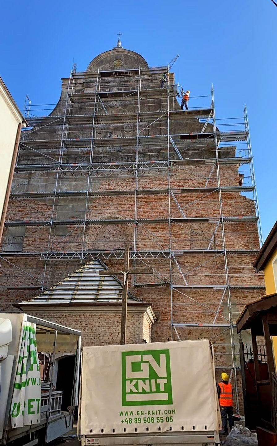 Renowacja zabytkowego kościoła z cegły, widoczne rusztowania, pracownicy w kaskach i kamizelkach odblaskowych, oraz ciężarówka z logo firmy budowlanej GN-KNIT.