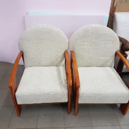 Dwa identyczne, odnowione fotele z jasną tapicerką i drewnianymi, lakierowanymi podłokietnikami, ustawione obok siebie na szarych płytkach.