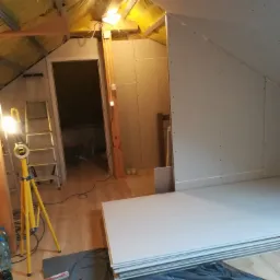 Poddasze w trakcie zabudowy płytami gipsowo-kartonowymi, widoczny stelaż, oświetlenie robocze i ułożone płyty na podłodze zabezpieczonej folią.