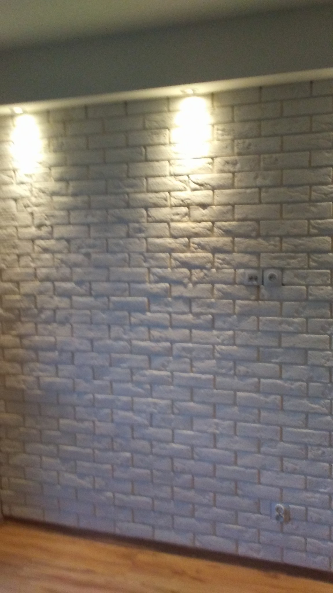 Ściana z białej cegły dekoracyjnej z widocznymi gniazdkami elektrycznymi i oświetleniem punktowym od góry.