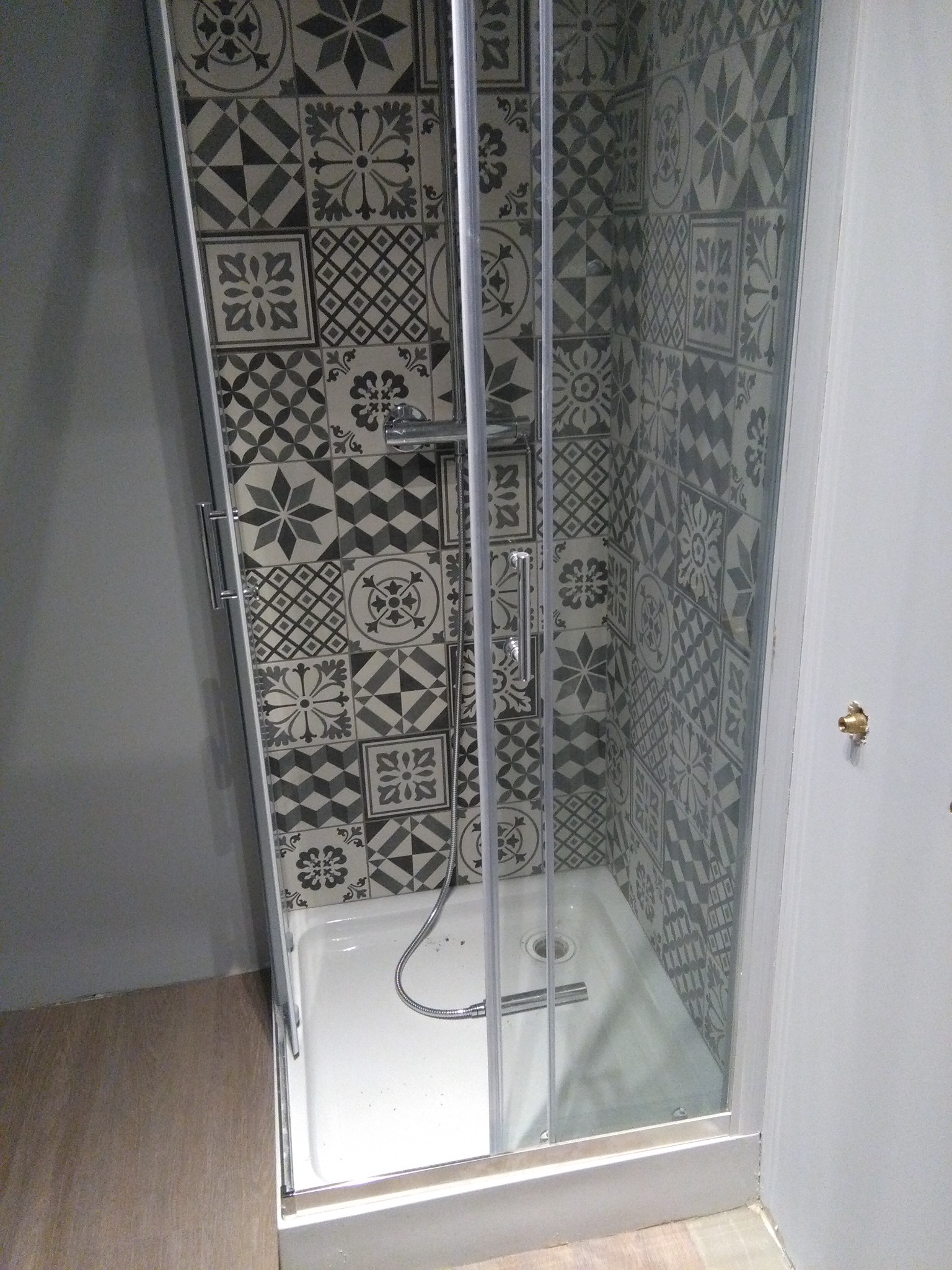 Kabina prysznicowa z wzorzystymi płytkami w odcieniach szarości, widoczny odpływ i słuchawka prysznicowa, jasne fugi, zamontowana na białym brodziku.