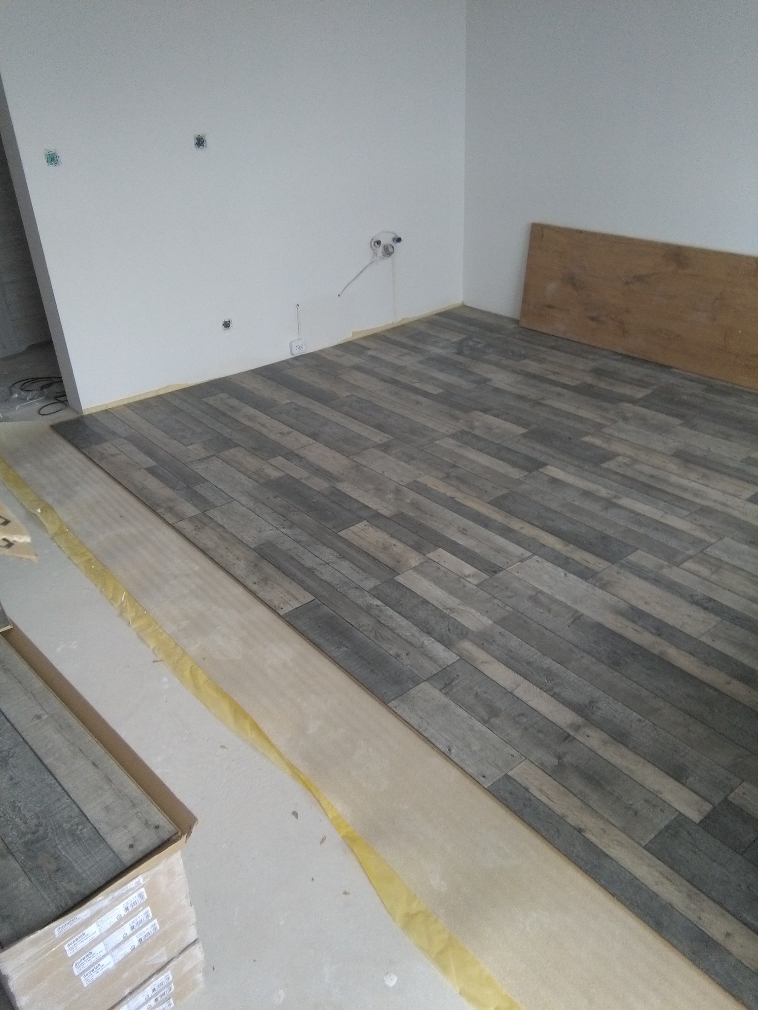 Układanie paneli podłogowych imitujących drewno w jasnym pomieszczeniu. Częściowo ułożona podłoga, widoczne kartony z panelami i taśma zabezpieczająca.