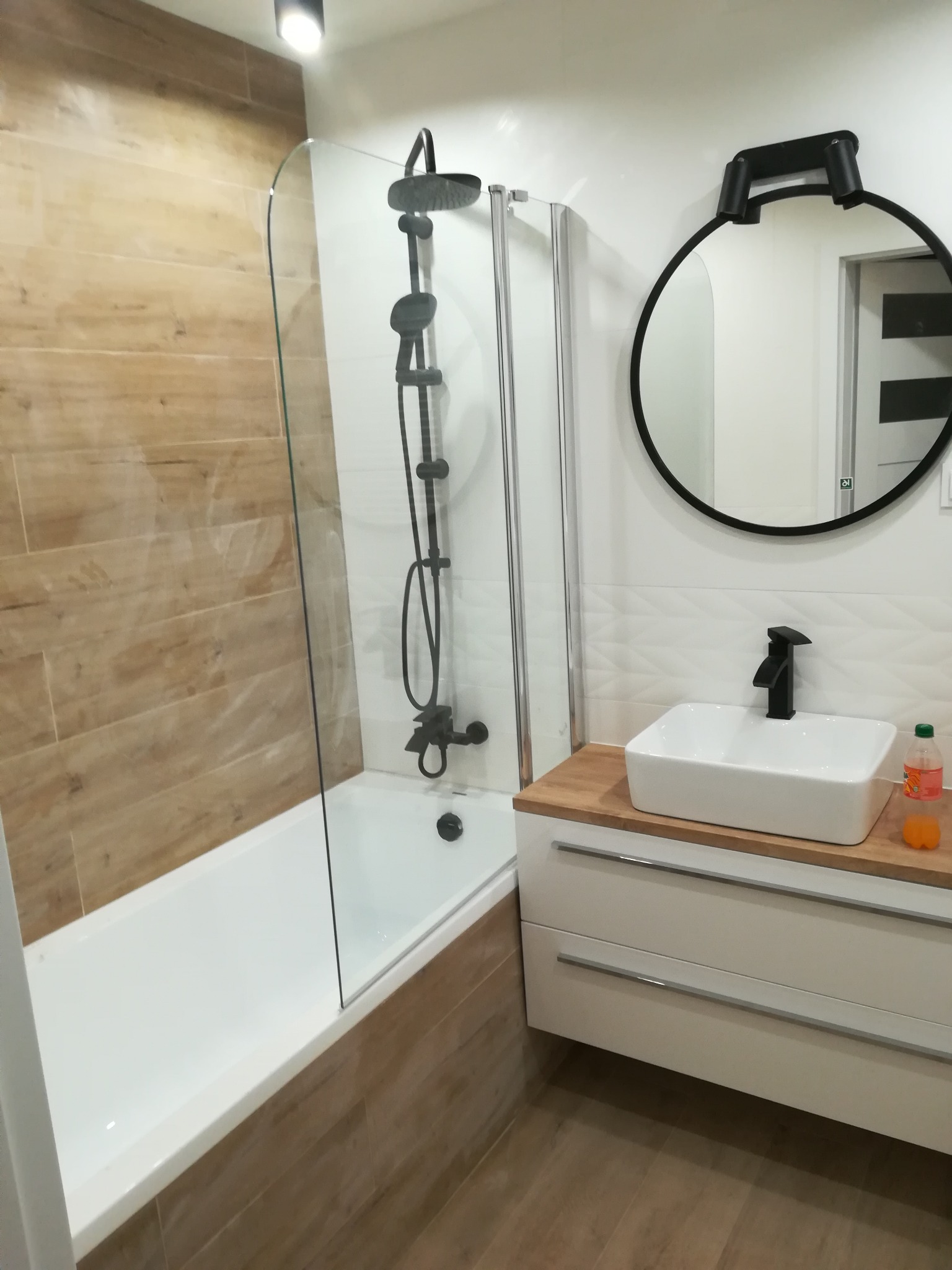 Nowoczesna łazienka z wanną obudowaną płytkami imitującymi drewno, kabiną prysznicową z czarnym zestawem natryskowym, białą umywalką nablatową na drewnianym blacie i okrągłym lustrem w czarnej ramie.