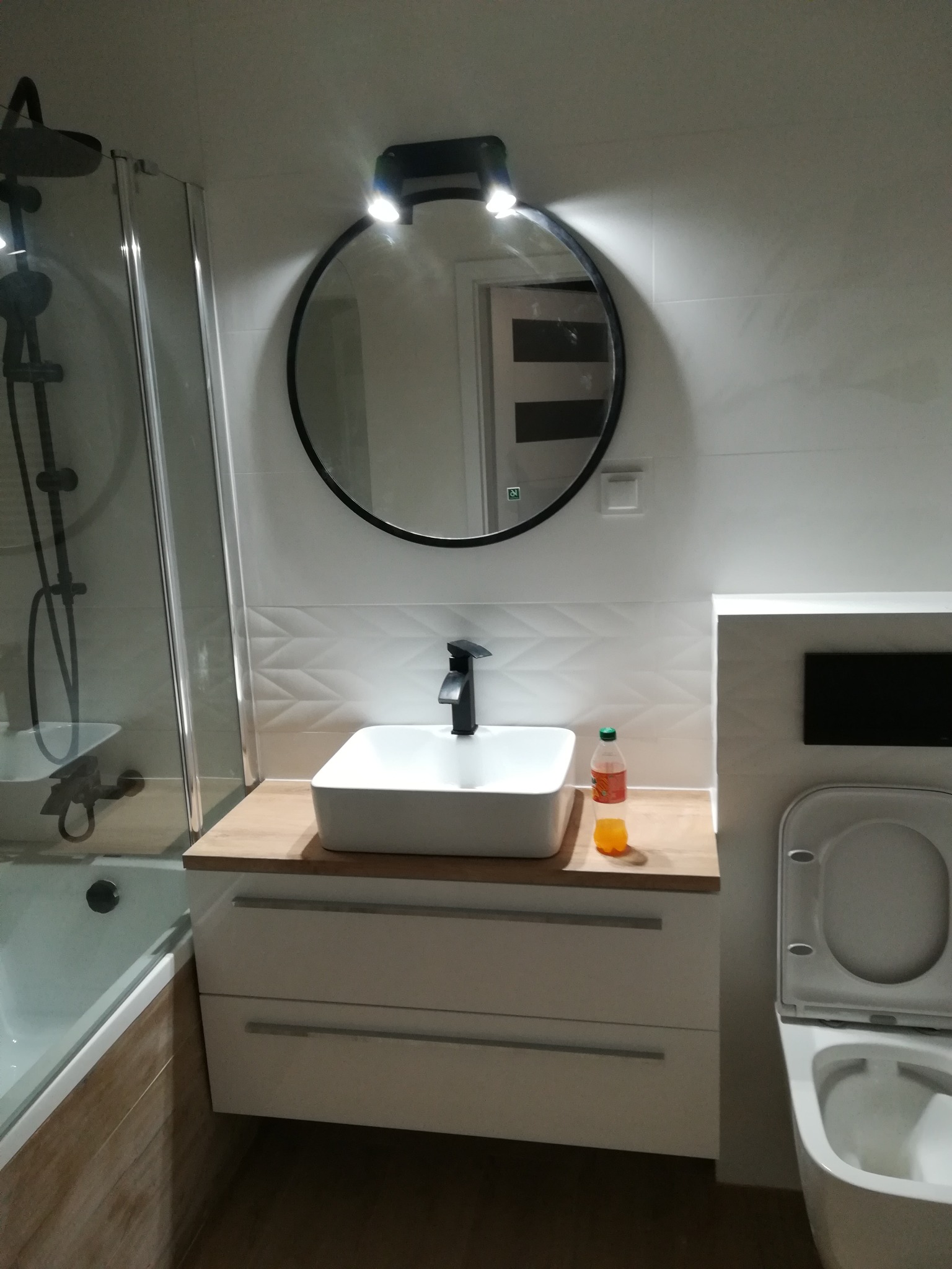 Nowoczesna łazienka z białą umywalką nablatową, czarną baterią i okrągłym lustrem z podświetleniem LED. Widoczna również wanna z parawanem nawannowym i toaleta. Na blacie stoi butelka napoju.