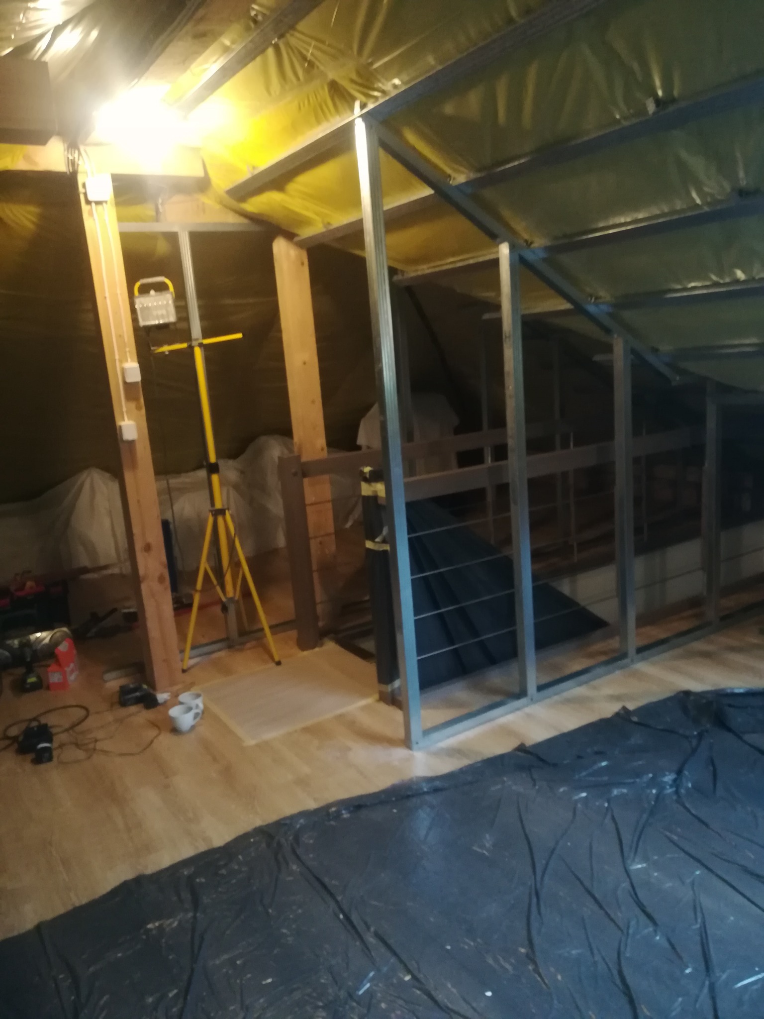 Stelaż z profili metalowych w trakcie montażu na poddaszu, widoczne ocieplenie folią, lampa robocza na trójnogu oraz rozłożona folia ochronna na podłodze.