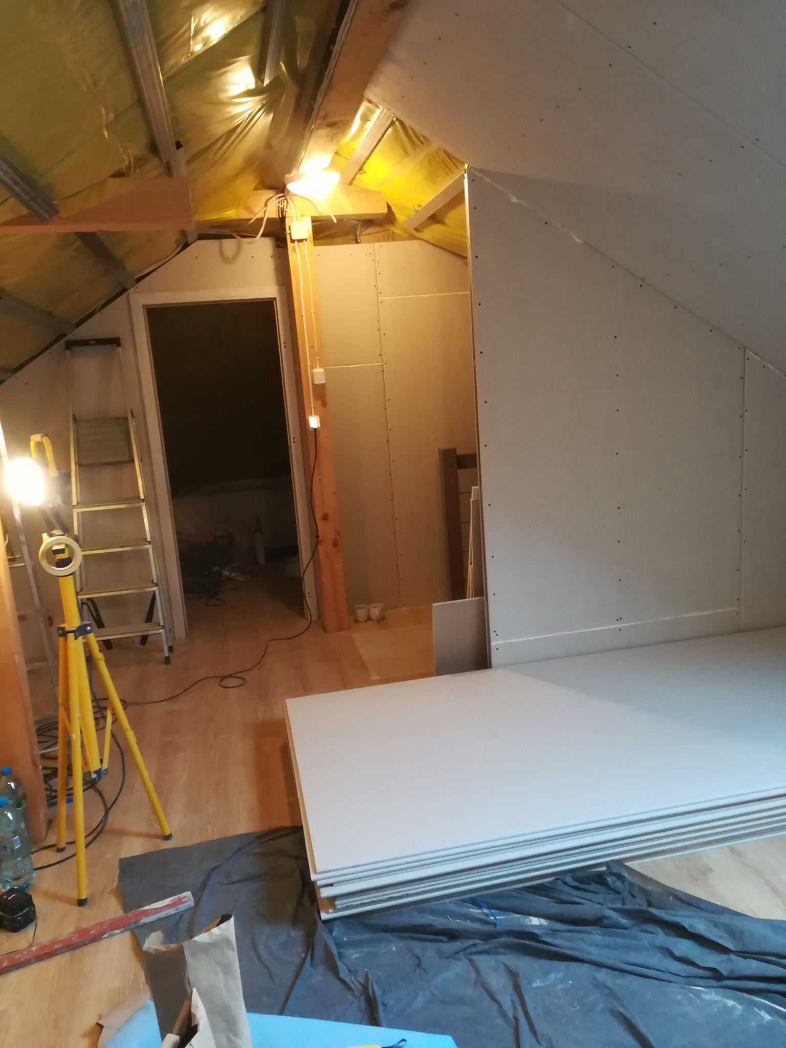 Poddasze w trakcie zabudowy płytami gipsowo-kartonowymi, widoczny stelaż, oświetlenie robocze i ułożone płyty na podłodze zabezpieczonej folią.